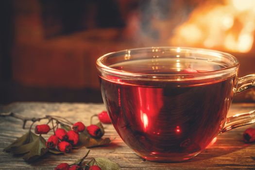 Tea with hawthorn in a glass cup on a wooden table in a room with a burning fireplace.