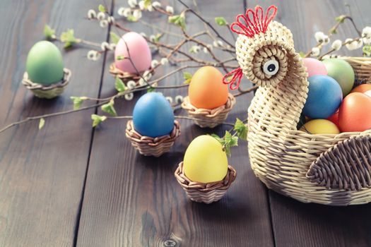 Easter composition - a wicker basket in the form of a chicken with colored eggs on a dark wooden table with willow twigs, place for text, copy space.