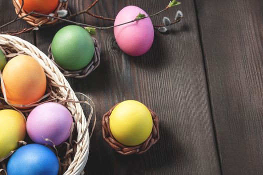 Easter composition - several colored eggs in a basket and on a dark wooden table with willow twigs, place for text, copy space.