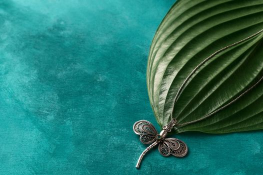 Silver dragonfly pendant on turcuoise background. The chain of pendant lies on big plant leaf. Perfect jewellery photo.