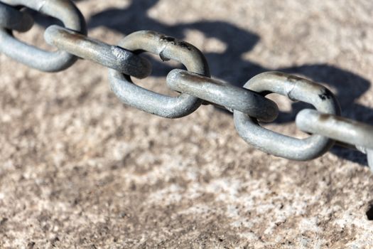 A light gray metal chain made of steel and showing its age is seen up close as it hangs just above textured concrete. It casts a shadow right below and behind it.