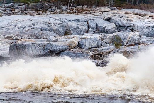 Water rushes into a river just below a hydroelectric dam. The whitewater crashes into the surface of the waterway creating mist and large white rapids. A rocky stone shore is seen behind the action.