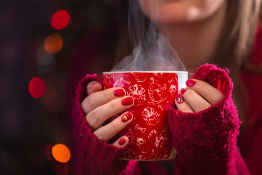 Detail of woman hands holding warm a cup of tea with steam. Blur Christmas background.