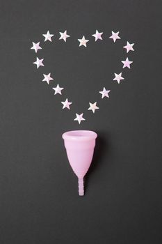 Pink menstrual cup on black background with heart, copy spase. Concept possibility of comfort use of cup at night, zero waste, savings, minimalism. Feminine hygiene product, flat lay. Vertical.