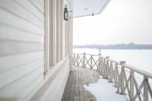 white snow on the wooden vintage open deck, old peeling paint light milk color, steel and copper lanterns hanging on the wall, snowy winter, snow lies on the river and the far shore of the earth is vi