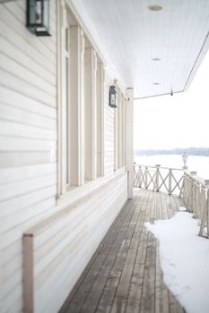 white snow on the wooden vintage open deck, old peeling paint light milk color, steel and copper lanterns hanging on the wall, snowy winter, snow lies on the river and the far shore of the earth is vi