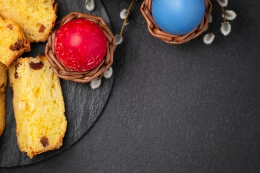 Painted Easter eggs in wicker coasters, pieces of Easter cake on a cutting board on a dark table - traditional Easter breakfast, top view, flat lay.