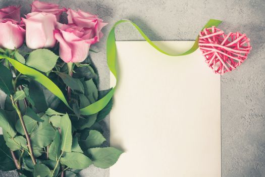 Flower arrangement - a bouquet of pink roses, a wicker heart and an empty sheet for inscription on a concrete surface, template for design or greeting card, top view, flat lay.