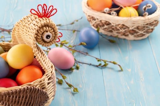 Wicker basket in the form of a chicken with colorful Easter eggs on blue wooden table - easter composition with copyspace.