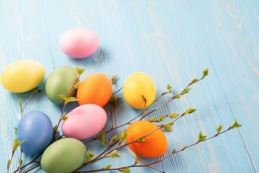 Multi-colored Easter eggs on a blue wooden table with twigs of birch with young leaves - Easter composition with copy space.