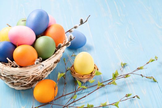 Multi-colored Easter eggs in a wicker basket on a blue wooden table with twigs of birch with young leaves - Easter composition with copy space.