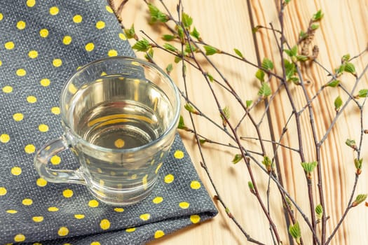 Birch juice on kitchen towel on a table in a glass mug, next to a branch of birch with young leaves.