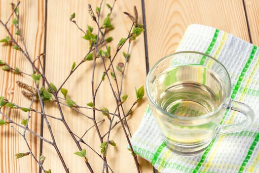 Birch juice on kitchen towel on a wooden table in a glass mug, next to a branch of birch with young leaves.