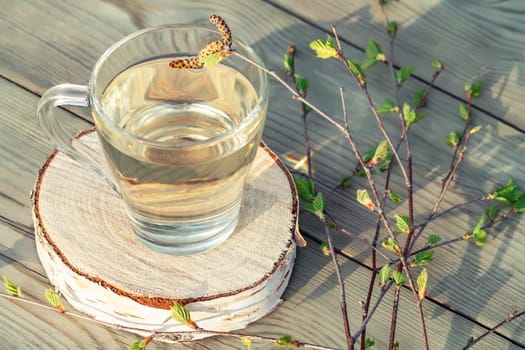 Birch juice on a wooden table in a glass mug, next to a branch of birch with young leaves.