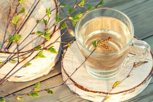 Birch juice on a wooden table in a glass mug, next to a branch of birch with young leaves.