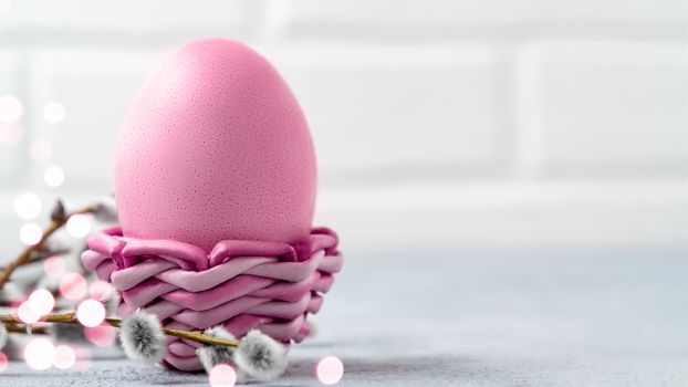 Pink Easter egg in a wicker stand on a gray table with pussy-willow twigs - Easter composition with copy space, horizontal banner.