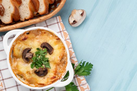 Casserole with chicken, mushrooms and cheese known in Russia as julienne in white bowl with herbs on a blue table, top view, flat lay, copy space.