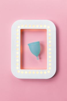 Turquoise menstrual cup in white luminous photo frame on pastel pink background. Concept zero waste living, savings, minimalism, these days. Feminine hygiene product, flat lay. Vertical orientation.