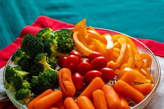 A glass dish of raw veggies consists of broccoli florets, baby carrots, grape tomatoes and sliced yellow bell peppers. The snack tray presents the vegetables on a table with a mat and tablecloth.