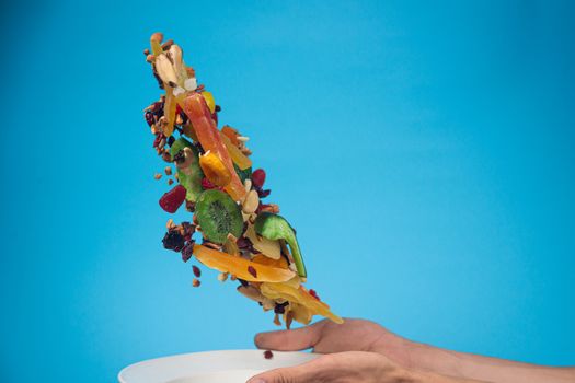 Male hands holding an empty white bowl on blue background. Candied fruits and nuts flying above the bowl. Stock photo of nutrient and healty food. Conceptual photo of vegan and vegetarial food and meal.