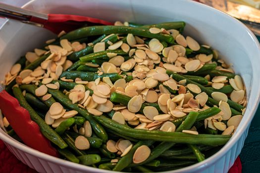 A white ceramic dish serves a side of green beans amandine, meaning with a garnish of almonds. The dish has French string beans and slices of almonds; tongs with red silicone grips are on the side.