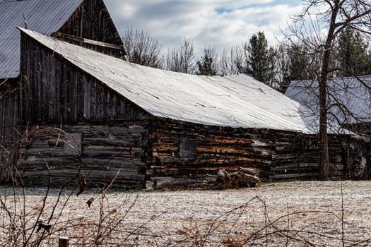 An old wooden log barn in the wintertime has a fresh coat of snow on its roof and surrounding grass. The snow continues to lightly fall around it.