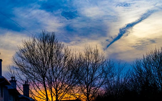 A vibrant, partly cloudy and textured sky is seen from a suburban backyard against the silhouettes of bare trees and roofs at sunset.