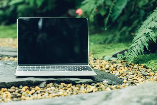 Laptop computer mockup has empty screen outside on stone in garden of coffee shop.