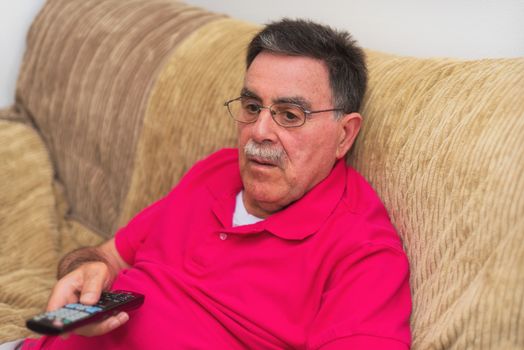 Senior man with remote control watching Tv on sofa at home