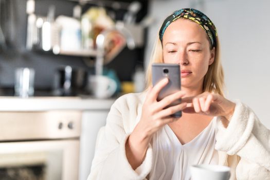 Beautiful caucasian woman at home, feeling comfortable wearing white bathrobe, taking some time to herself, drinking morning coffee and reading news on mobile phone device in the morning.