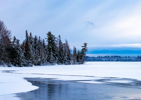 Snow-covered pine trees cover a small peninsula of woodland that extends out into a lake. Much of the lake is now frozen in winter, with liquid water seen in the foreground.