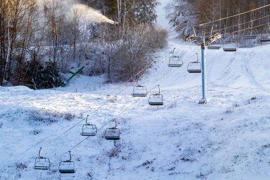 A chairlift running up the slope of a ski hill is standing motionless with its seats hanging from it. The hill below it needs more snow, and a snow machine prepares the ski hill for use behind it.