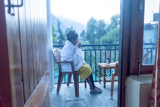 Young freezing tourist Traveler woman wraps up in winter clothing sitting on holiday villa balcony, holding a hot cup of tea in morning. Travel vacation active life and healthy lifestyles concept. Mountain city on background.