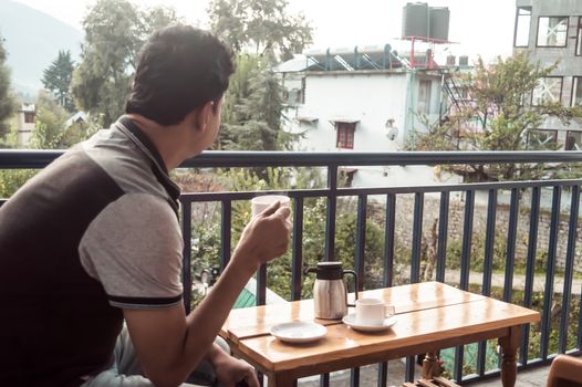 Mature adult tourist Solo business traveller of Indian ethnicity in casual clothing sitting on balcony of a holiday villa and holding a hot cup of tea in morning on Mountain city background. Travel vacation active life and healthy lifestyles concept.
