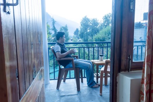 Mature adult tourist Solo business traveller of Indian ethnicity in winter clothing sitting on balcony of a holiday villa and holding a hot cup of tea in morning on Mountain city background. Travel vacation active life and healthy lifestyles concept.