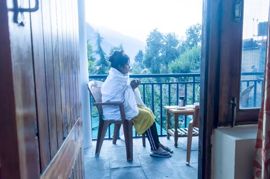 Young freezing tourist Traveler woman wraps up in winter clothing sitting on holiday villa balcony, holding a hot cup of tea in morning. Travel vacation active life and healthy lifestyles concept. Mountain city on background.