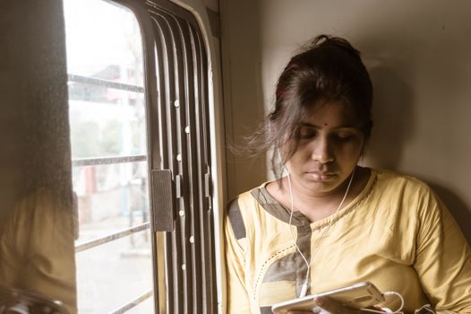 Woman using mobile phone while traveling solo in passenger train. Traveler enjoying Modern technology on the move in everyday life and travel. Close up portrait - Young Adult Lady - Indian ethnicity.