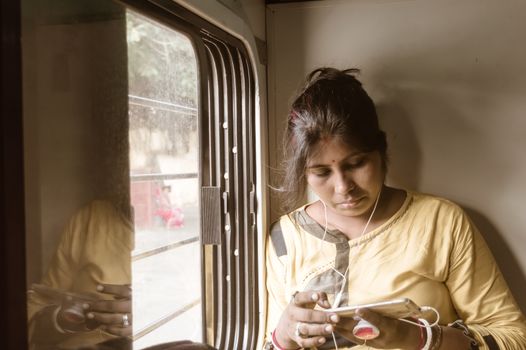 Woman using mobile phone while traveling solo in passenger train. Traveler enjoying Modern technology on the move in everyday life and travel. Close up portrait - Young Adult Lady - Indian ethnicity.