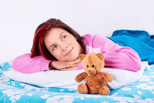 Beautiful young girl in pink pajamas lying in bed with her cute plush teddy bear daydreaming of something nice.