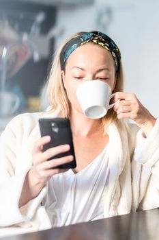 Beautiful caucasian woman at home, feeling comfortable wearing white bathrobe, taking some time to herself, drinking morning coffee and reading news on mobile phone device in the morning.