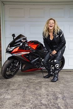 blond woman wearing motocyclist leather clothes, laughing hard in from of a sport motocycle