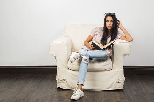 young woman sitting on a couch, with her glasses in her hair, reading a hard cover book