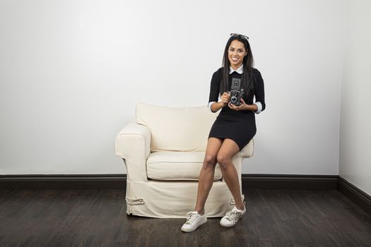 Young woman, with happy expression, using a vintage camera, sitting on the arm of a couch