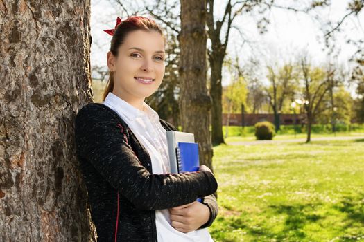 Beautiful casual student girl holding notebooks leaning against a tree in the park. Student taking a break from studying in the park.