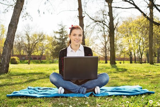 Beautiful casual student girl with laptop outdoors. Smiling woman in the park sitting on a blanket with computer, surfing the Internet or preparing for exams. Technology, education and remote working concept.