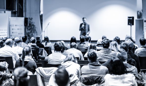Male public speaker giving a talk at business meeting conference. Rear view of unrecognized people sitting in audience in the conference hall. Business and entrepreneurship concept. Blue toned.