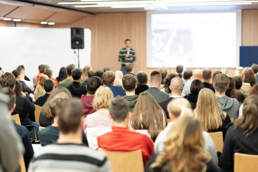 Speaker giving presentation in lecture hall at university. Participants listening to lecture and making notes.
