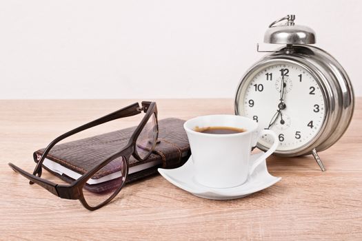 Close up of a leather covered planner notebook, glasses, alarm clock and a cup of coffee placed on a wooden table.