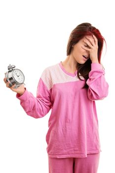 Being late concept. Portrait of a young woman in pink pajamas holding an alarm clock and covering her face with her hand, looking tired and annoyed, isolated on white background. Girl panicking about being late.
