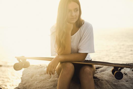 Beautiful and fashion young woman posing at the sunset with a skateboard